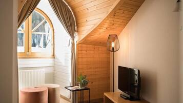 A cozy room with a sloped ceiling, a television, a small table with a plant, and two ottomans.