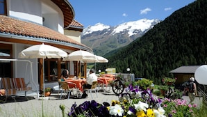 A hotel patio with tables and chairs, surrounded by colorful flowers and a mountain backdrop. - Hotel Alpenjuwel (Curon Venosta)