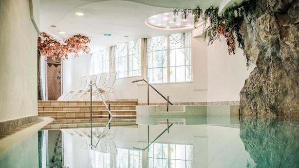 A modern indoor pool with clear glass walls, a central staircase, and natural decor elements. - Hotel Alpenjuwel (Curon Venosta)