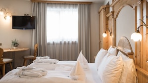 A hotel room with two beds, a TV, a desk, and a large window with curtains. - Hotel Alpenjuwel (Curon Venosta)