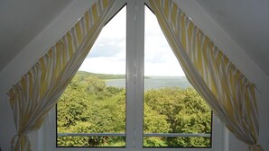 A window with yellow curtains, offering a view of a lush green landscape and a body of water. - Hotel Sonnenhaken (Buschvitz)