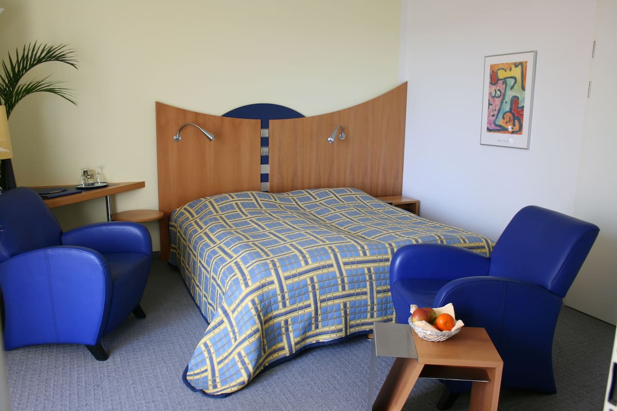 Une chambre d’hôtel avec un lit, deux fauteuils bleus, une petite table sur laquelle se trouve un panier de fruits, un bureau avec une lampe et une œuvre d’art murale.