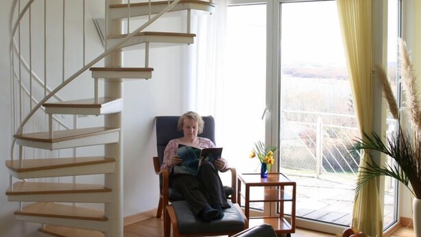 A spacious living area with a wooden staircase, a comfortable sofa, a small table with a vase, and a view through large windows. - Hotel Sonnenhaken (Buschvitz)