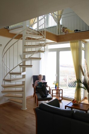 A spacious living area with a wooden staircase, a comfortable sofa, a small table with a vase, and a view through large windows. - Hotel Sonnenhaken (Buschvitz)