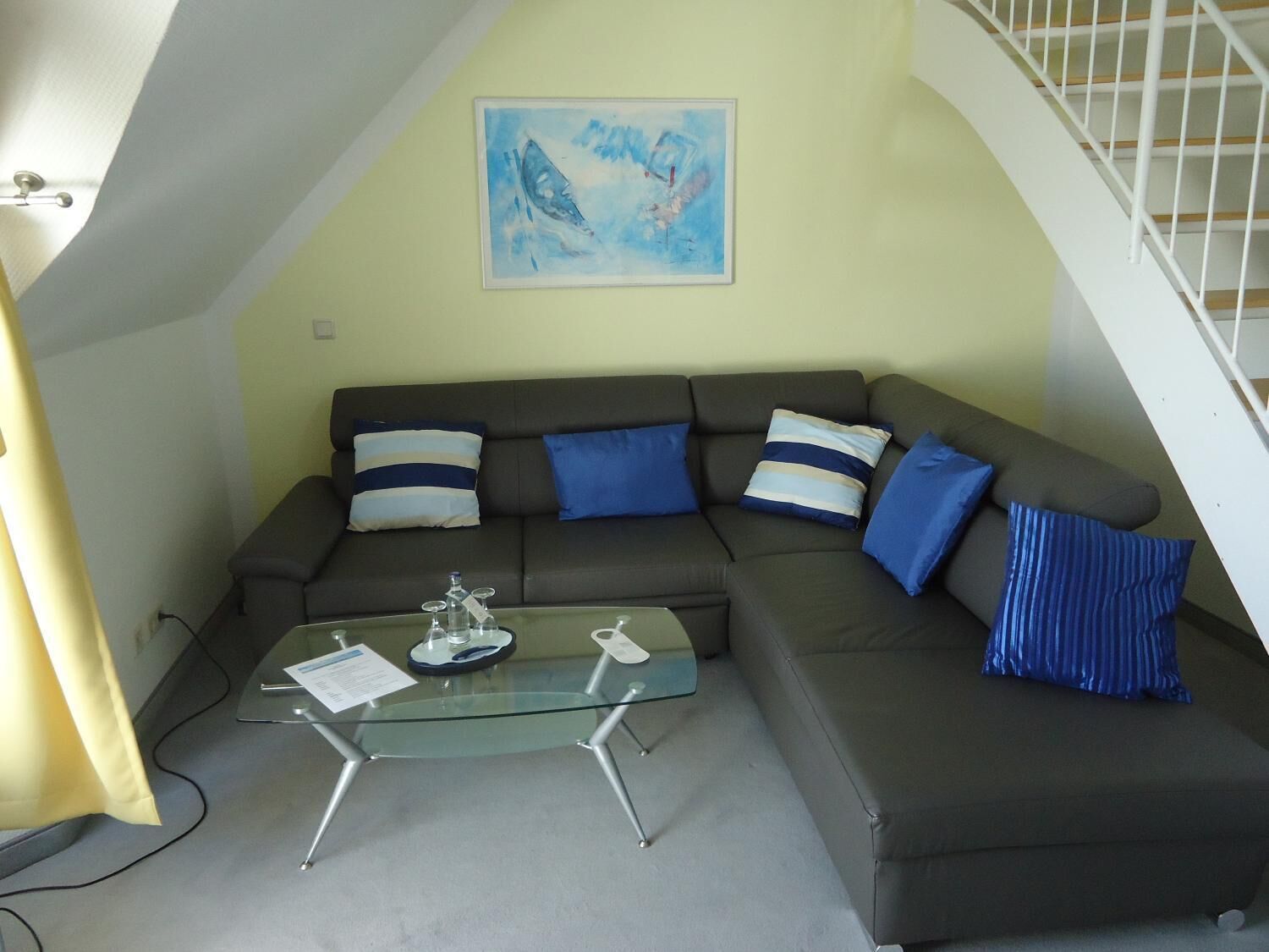 A modern living room with a dark grey sofa, blue and white striped pillows, a glass coffee table, and a painting on the wall.