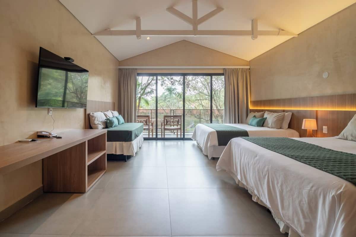 A hotel room with two beds, a desk, a TV, and a balcony with a view of greenery.