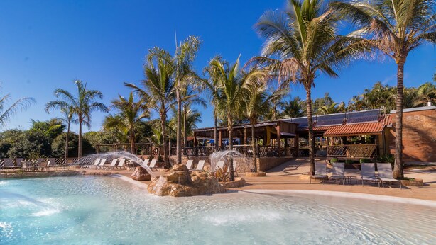 Et poolområde med klart vand, liggestole, palmer og en bygning med rødt tag.