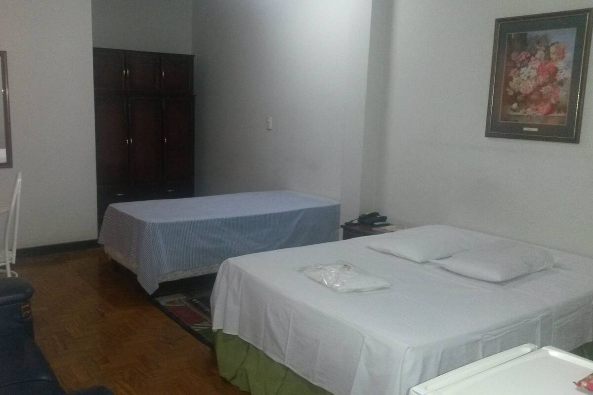 A small, dimly lit room with two single beds, a wooden wardrobe, a white chair, and a framed picture on the wall.