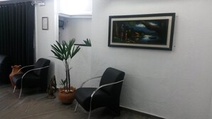 A waiting area with two black chairs, a potted plant, and a framed painting on the wall. - Hotel Fátima (Divinopolis)