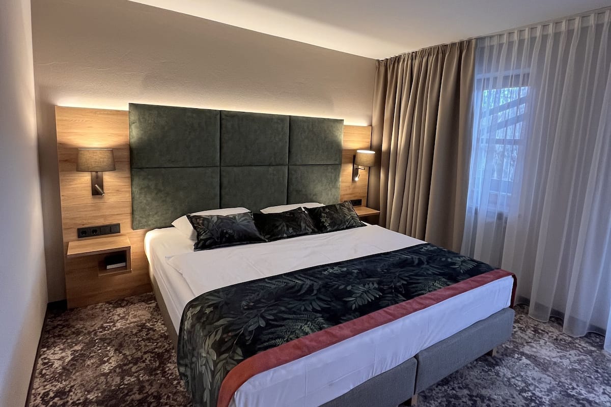 Une chambre d’hôtel avec un grand lit, deux lampes de chevet, une table de nuit en bois et une fenêtre avec des rideaux légers.