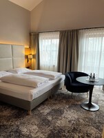 Habitación de hotel con una cama grande, una mesa redonda con dos botellas y vasos, una silla y una lámpara.