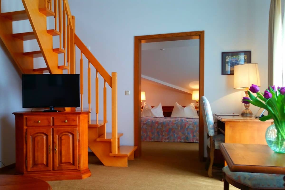 Une chambre d’hôtel avec un escalier en bois, un téléviseur posé sur une commode en bois, un lit avec une literie colorée, un bureau avec une lampe et un vase rempli de fleurs.