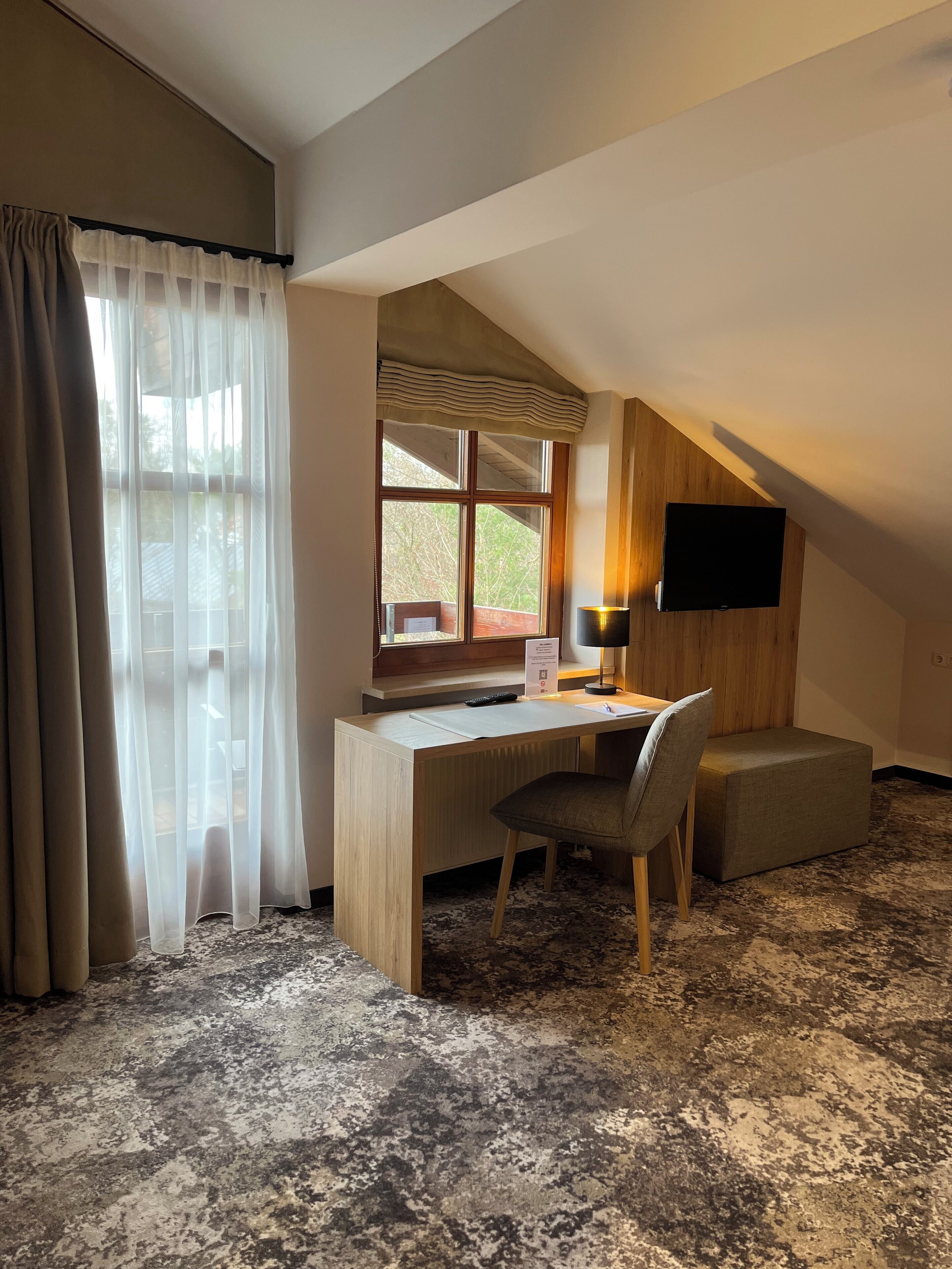 Una habitación con un escritorio de madera, una silla, un televisor montado en la pared y una ventana con cortinas.