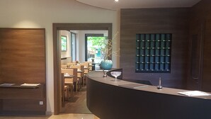 Un espace d’accueil moderne avec un comptoir en bois aux lignes courbes, un coin salon avec des tables et des chaises, et une vue sur l’extérieur.
