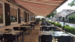 Outdoor dining area with tables and chairs under a striped awning, adjacent to the city.