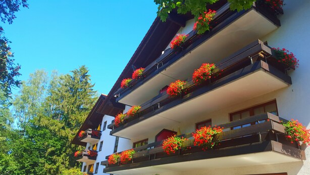 Un immeuble à plusieurs étages, doté de balcons ornés de fleurs rouges, et entouré d’arbres.