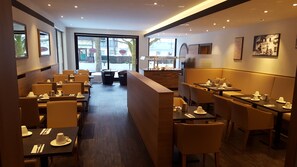 Enjoy dining in our spacious dining area with tables, chairs, and a reception desk, surrounded by large windows and artwork.