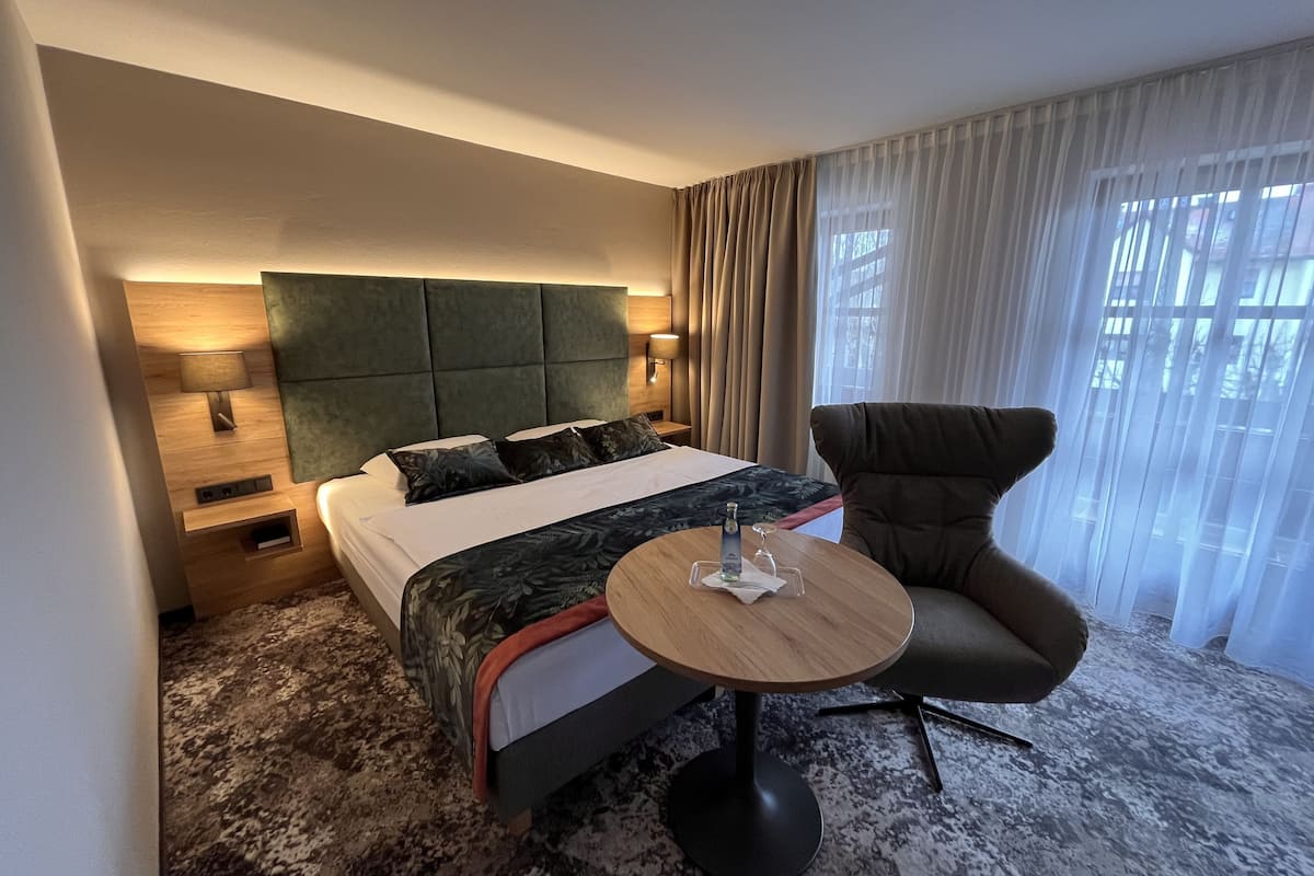 Une chambre d’hôtel moderne avec un grand lit, une table ronde, un fauteuil confortable et une vue sur l’extérieur à travers des rideaux légers.