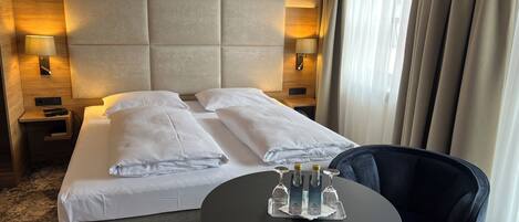 Una habitación de hotel con una cama grande, dos lámparas de noche, una mesita con botellas y una silla.