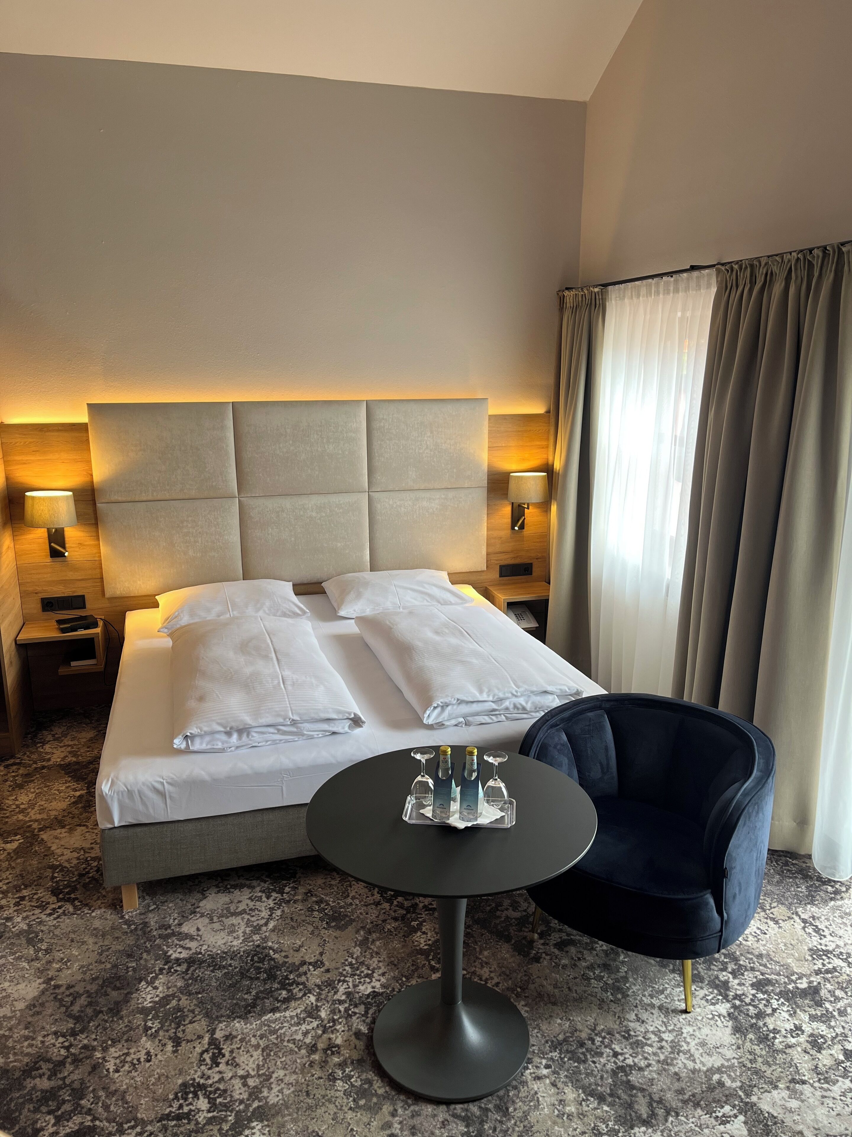 Habitación de hotel con cama, lámparas de noche, una mesa redonda con vasos y un sillón oscuro.