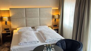 Habitación de hotel con cama, lámparas de noche, una mesa redonda con vasos y un sillón oscuro.