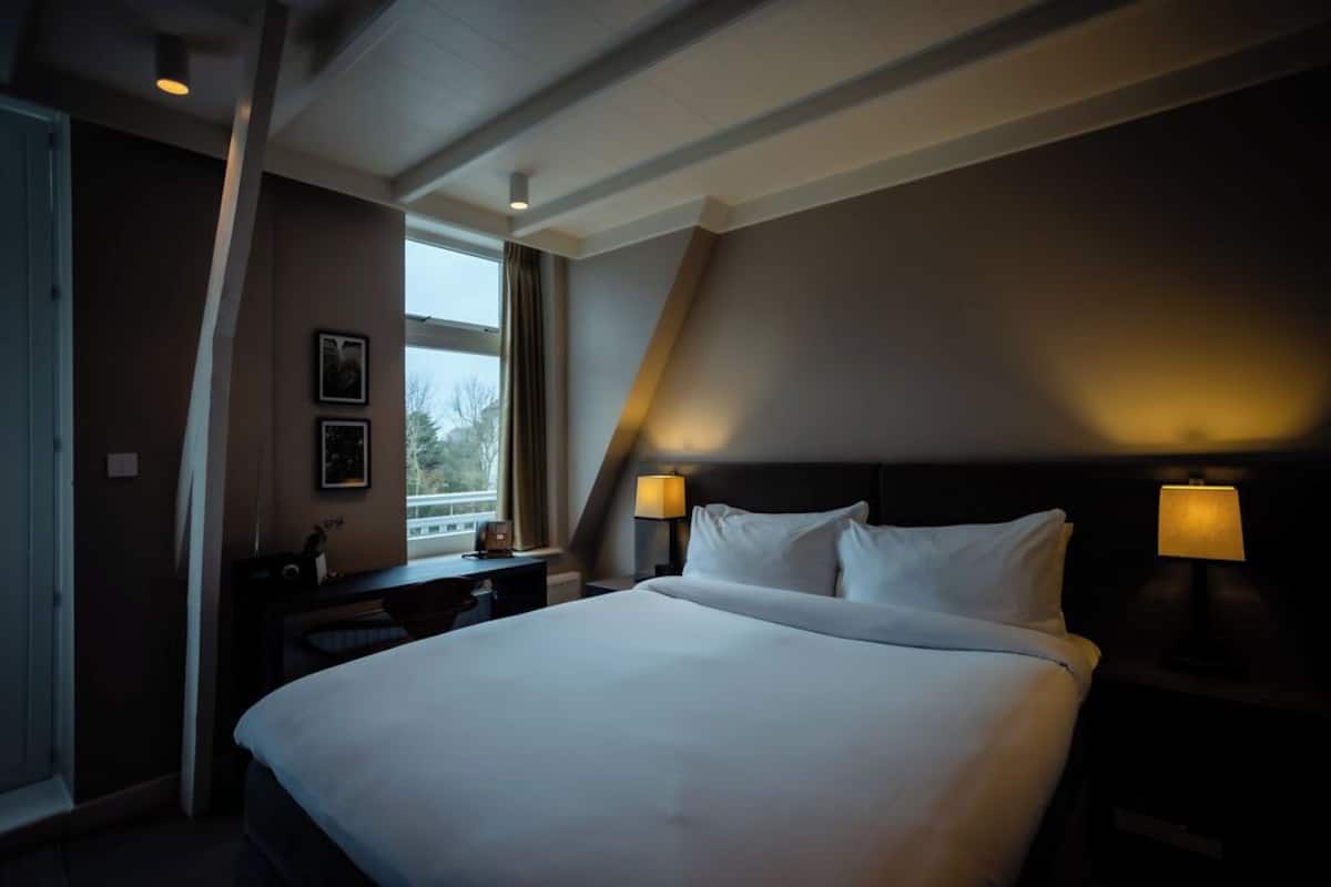 Une chambre d’hôtel avec un grand lit, deux tables de chevet avec des lampes, une fenêtre avec des rideaux et un tableau au mur.