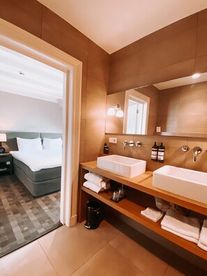 Une salle de bain d’hôtel moderne avec deux vasques, un grand miroir et une table de chevet avec une lampe.