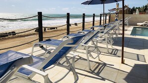 Zona de piscina frente al mar con sillones de descanso y sombrillas, una barrera de cuerda y vista al océano.
