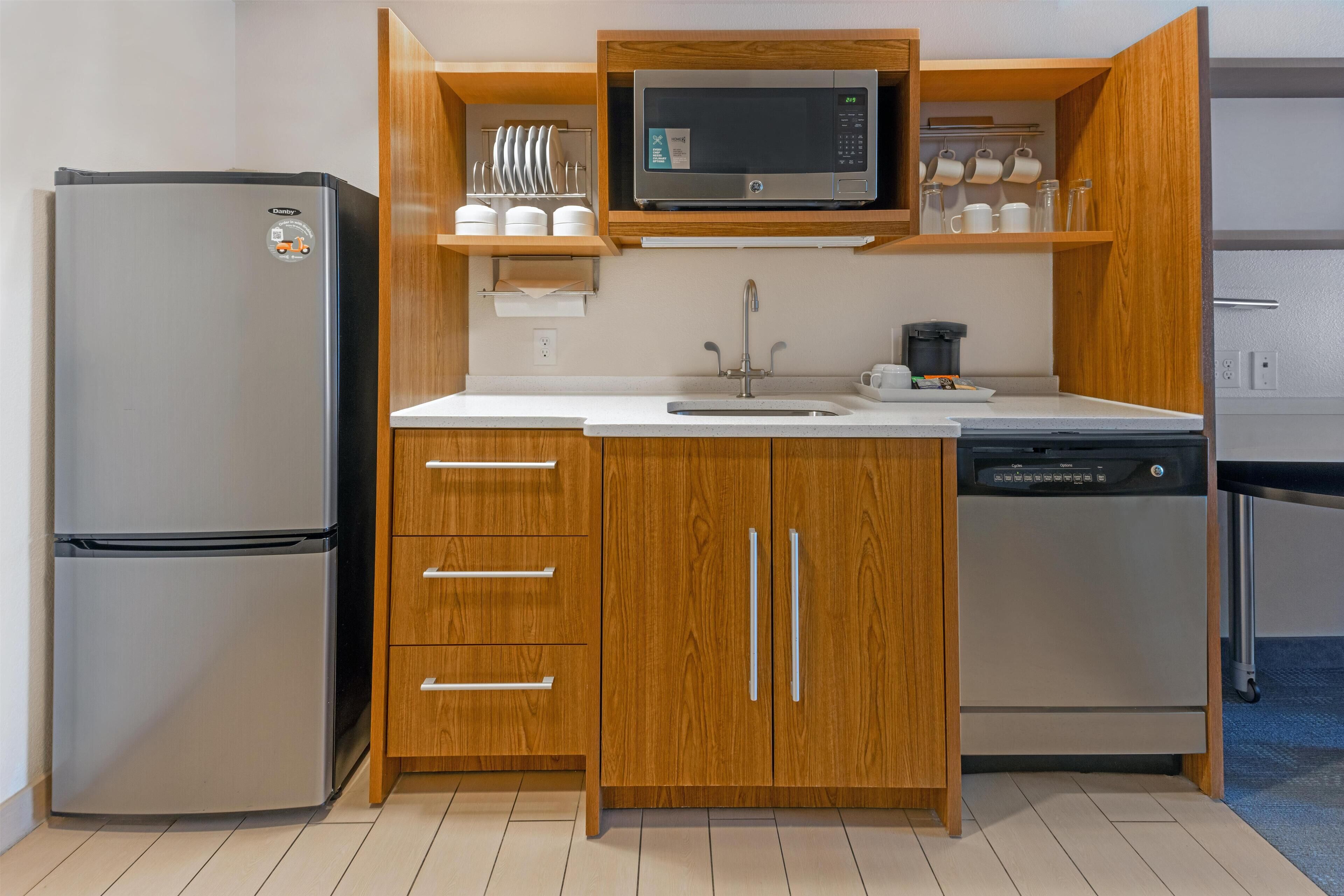 A compact kitchen with a stainless steel refrigerator, wooden cabinets, a microwave, and a dishwasher.