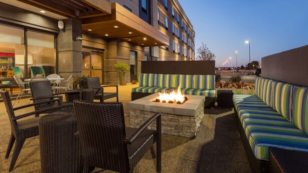 An outdoor seating area with wicker chairs and a fire pit, a striped sofa, and a stone fireplace. - Home2 Suites by Hilton Fargo, ND (Fargo)