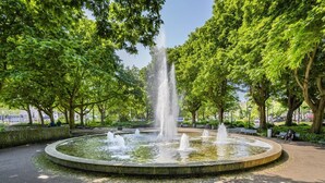 Une fontaine circulaire dotée de plusieurs jets d’eau, située dans un parc, entourée d’arbres et de bancs.