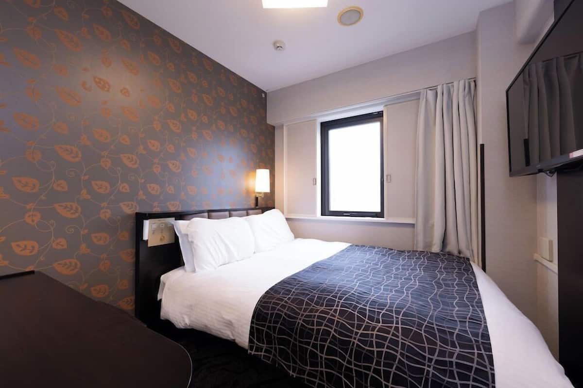 Ett hotellrum med en säng, ett skrivbord, ett fönster med gardiner och en tv på väggen.