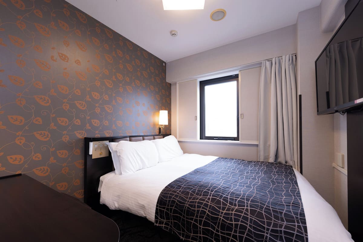 Ein Hotelzimmer mit Bett, Schreibtisch, Fenster mit Vorhängen, Fernseher und Wand mit Blättermotiv.