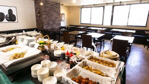 Een buffetopstelling met diverse gerechten, borden, tafels en stoelen, gerangschikt in een eetruimte met een bakstenen muur en ramen.