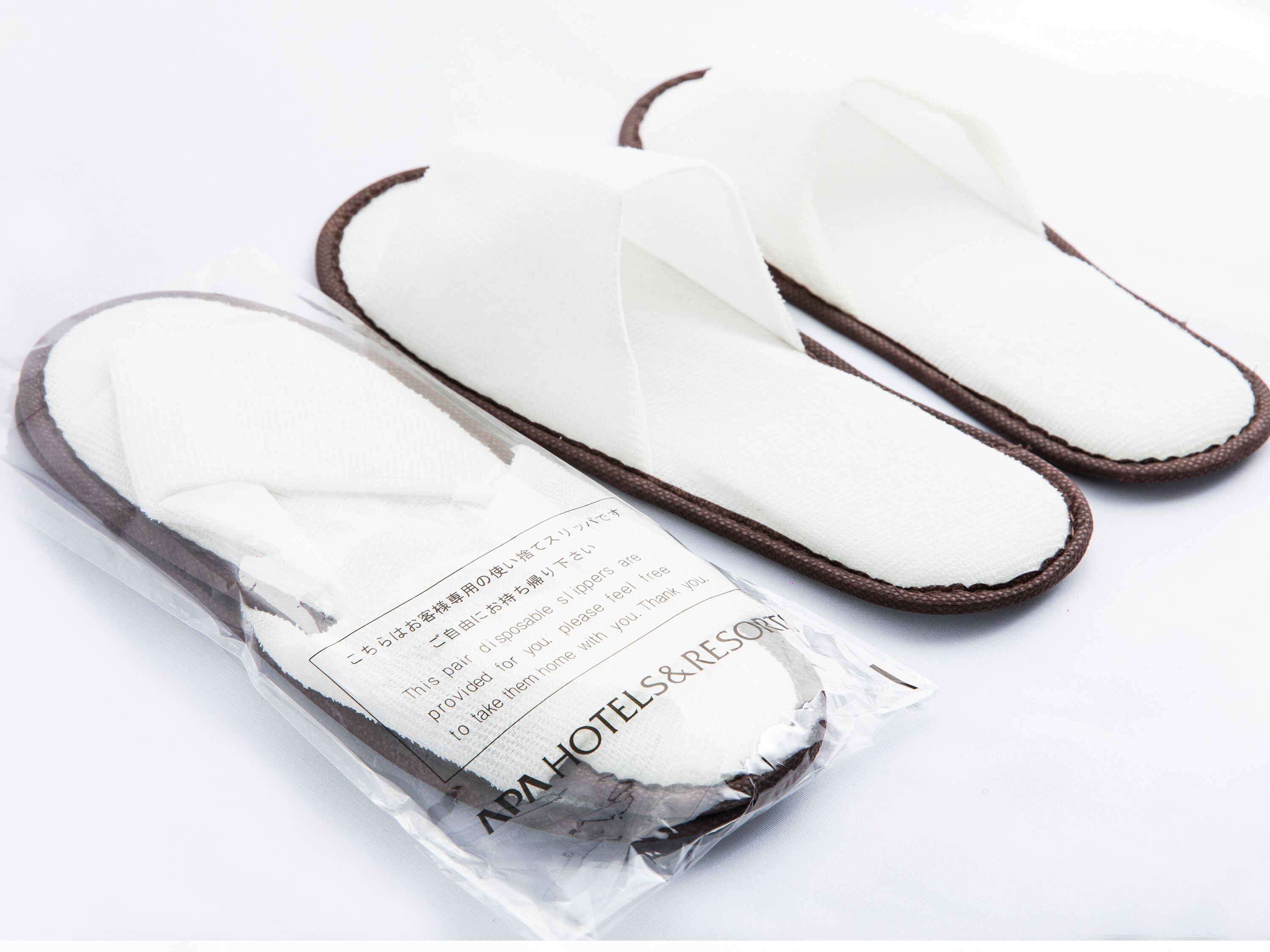 A pair of white slippers with brown trim, packaged in a clear plastic bag with a label.