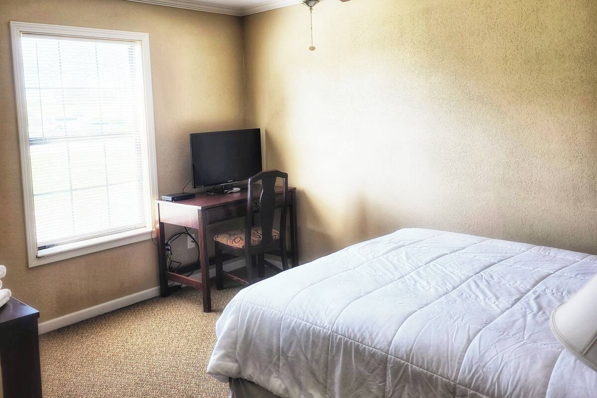 A bedroom with a bed, a desk with a computer, a ceiling fan, and a window.