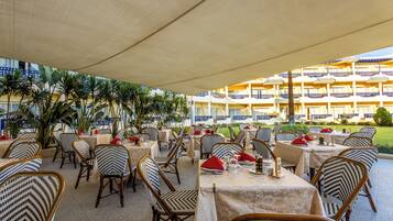 An outdoor dining area with tables and chairs, a covered section, and a building with balconies in the background.
