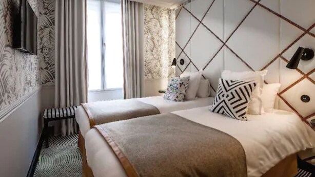A hotel room with two beds, a large window with curtains, a patterned wall, and a geometric headboard. - Hôtel Jardin le Bréa (Paris)