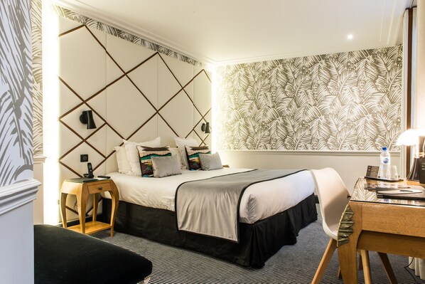 Hotellihuone, jossa on sänky, yöpöytä, tuoli, työpöytä ja kuviollinen tapetti.