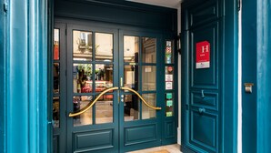 A teal double door with a brass handle, a red sign with a 'H' and three stars, and various stickers on the door. - Hôtel Jardin le Bréa (Paris)