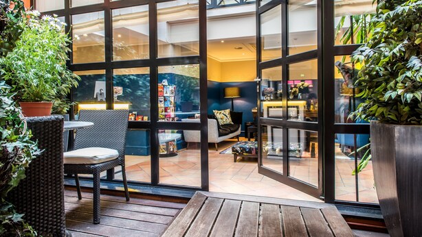A spacious indoor area with large glass doors leading to a living room with a sofa and a coffee table. There are potted plants and a wicker chair visible. - Hôtel Jardin le Bréa (Paris)
