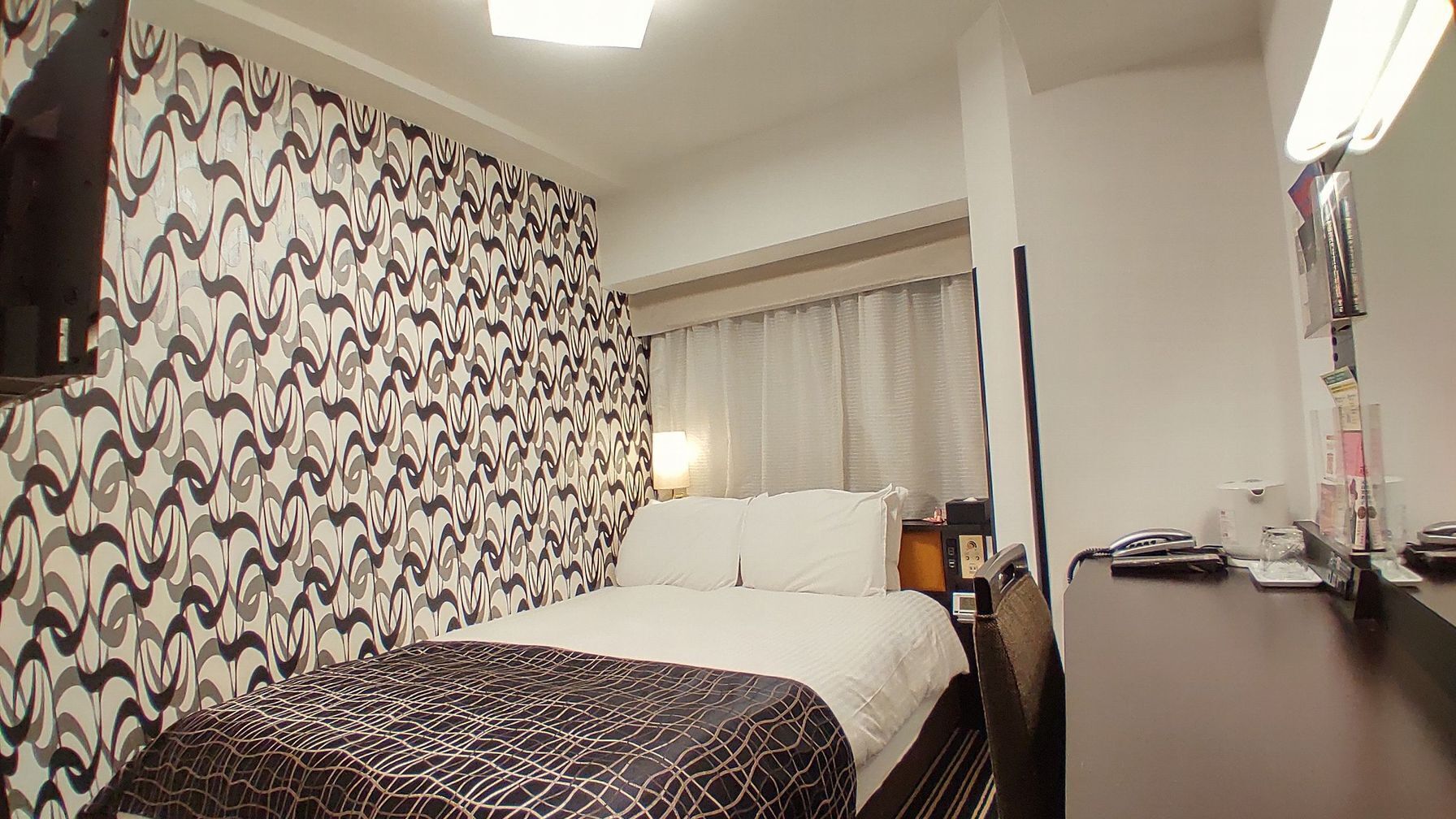 Ett hotellrum med en vägg som har ett mönster, en säng med ett mönstrat överkast, ett skrivbord med en telefon och ett nattduksbord med en lampa.