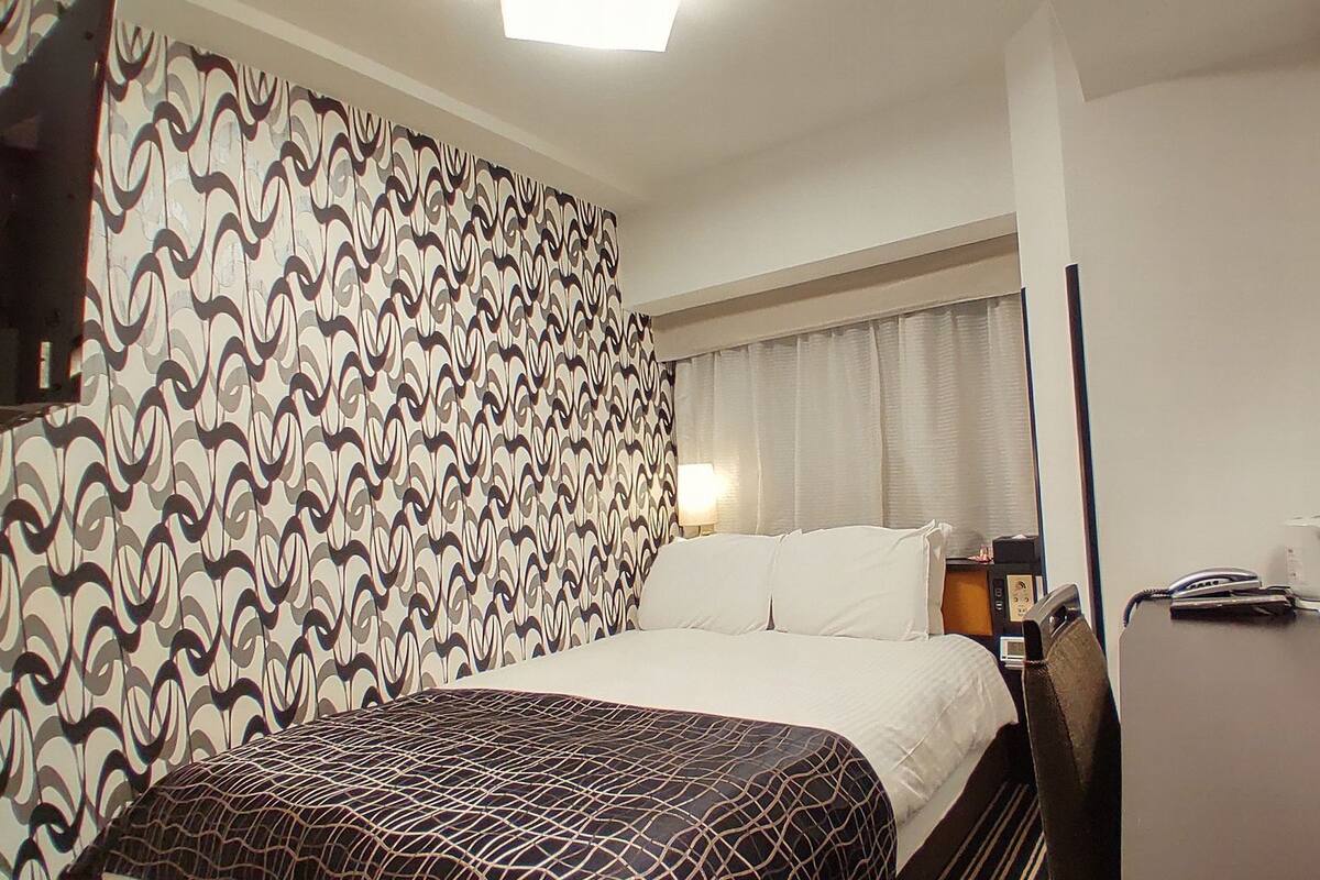 A hotel room with a patterned wall, a bed with a patterned bedspread, a desk with a telephone, and a nightstand with a lamp.
