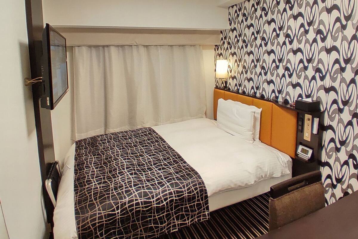 A hotel room with a bed, a bedside table, a lamp, a wall clock, and a patterned curtain.