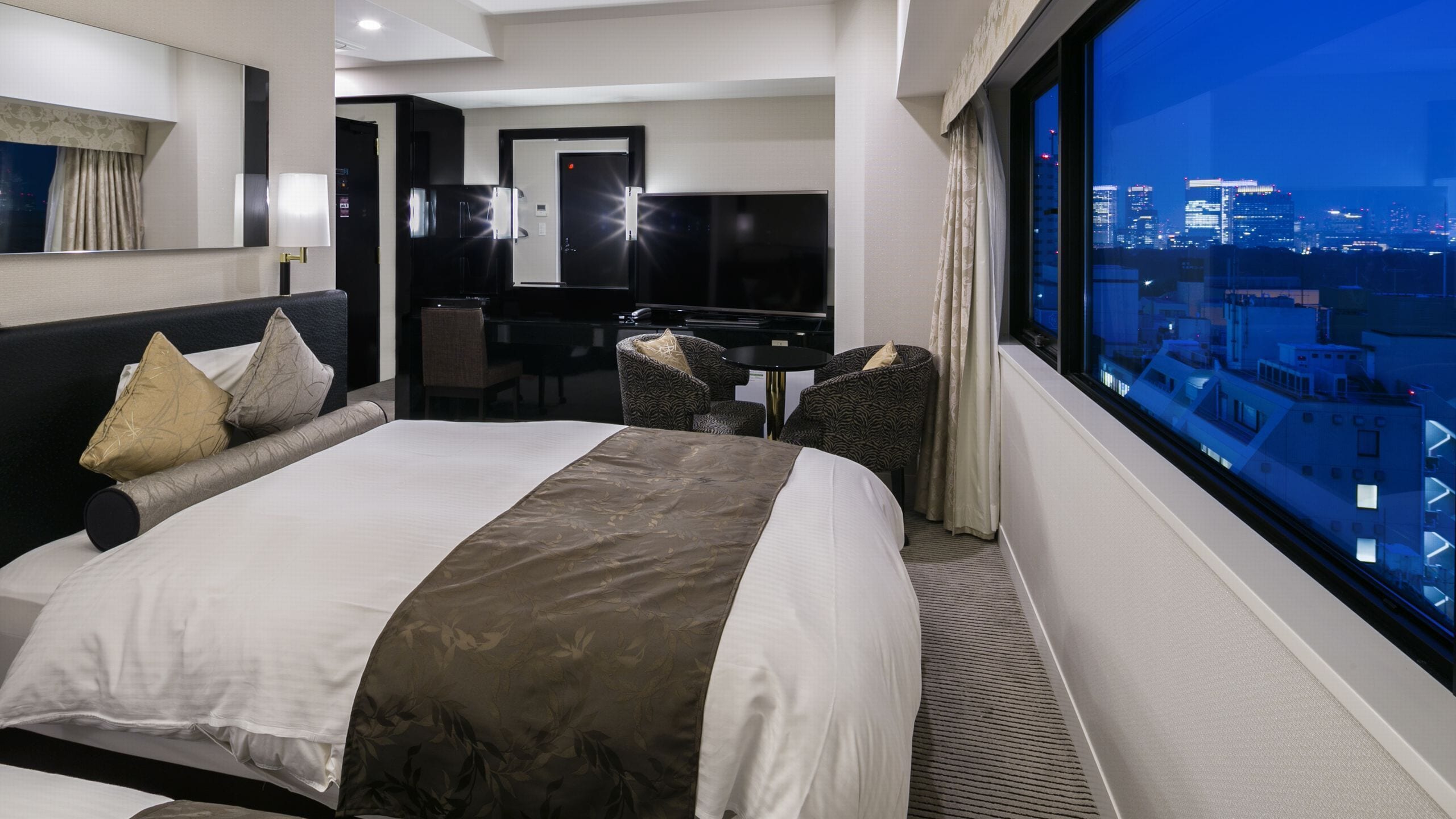 Ett hotellrum med en stor säng, ett skrivbord, en stol och utsikt över staden.