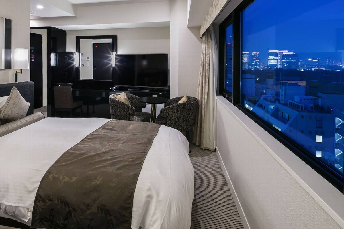 A hotel room with a large bed, a desk, a chair, and a view of the cityscape.