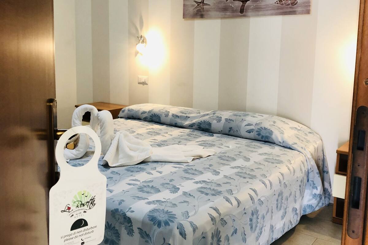 Une chambre d’hôtel avec un mur à rayures, un lit avec une literie à motifs floraux, un tableau représentant un arbre et un porte-serviettes avec un sac.