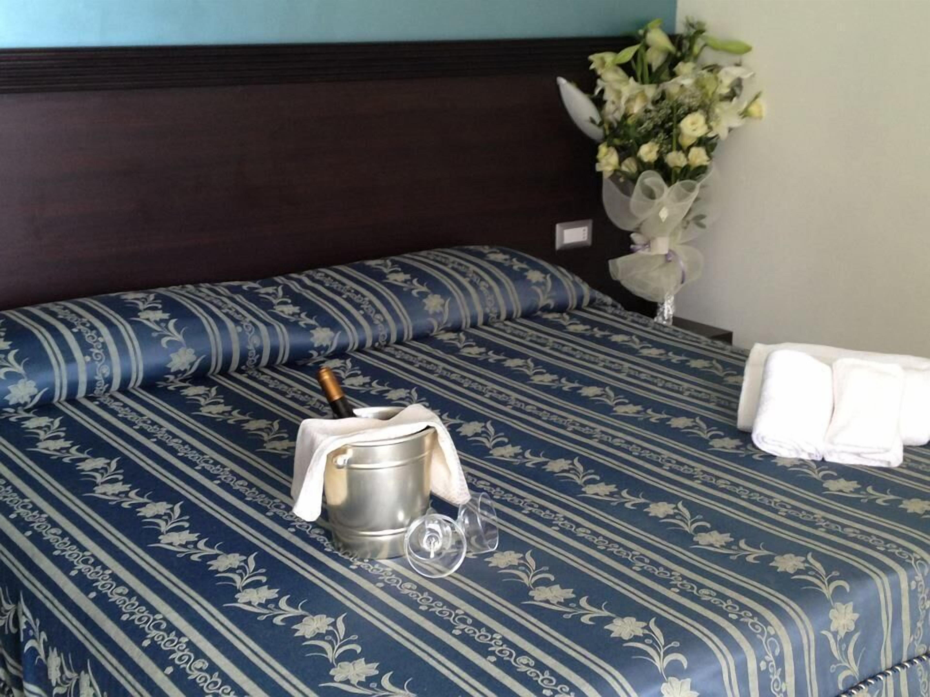 Habitación de hotel con una cama cubierta por una colcha azul estampada con motivos florales, un cubo metálico con una botella y un vaso. Hay dos toallas blancas sobre la cama.