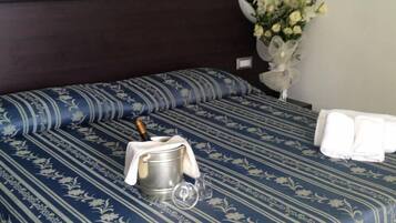 Habitación de hotel con una cama cubierta por una colcha azul estampada con motivos florales, un cubo metálico con una botella y un vaso. Hay dos toallas blancas sobre la cama.