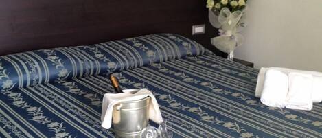 Habitación de hotel con una cama cubierta por una colcha azul estampada con motivos florales, un cubo metálico con una botella y un vaso. Hay dos toallas blancas sobre la cama.
